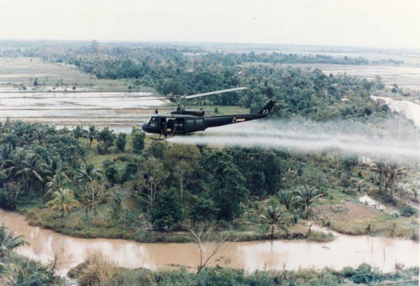 A helicopter in flight over rural Vietnam, leaving a trail of Agent Orange in its path.