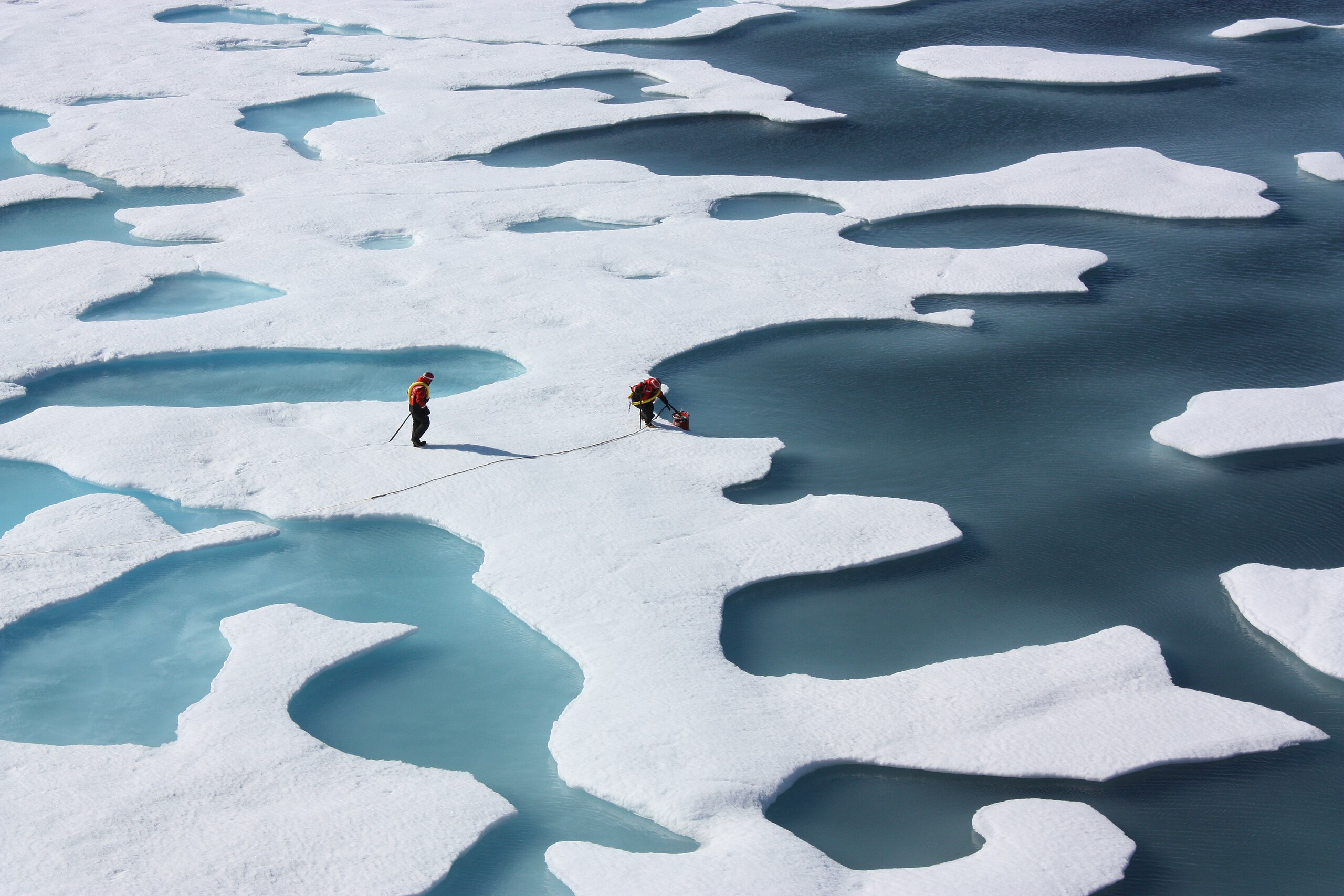 Two people shown from a distance stood on white ice, with several blue melt ponds around them.