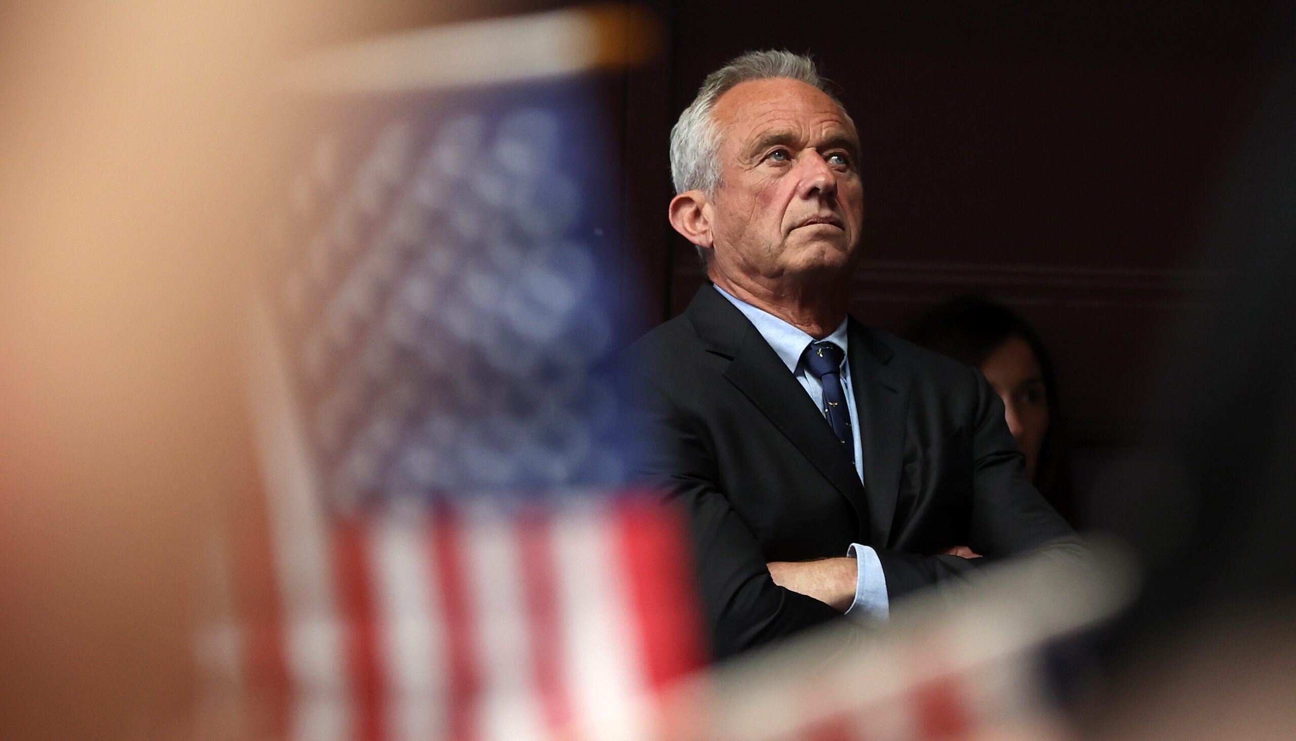 Robert F. Kennedy Jr. looks on with an out of focus United States flag in the foreground.