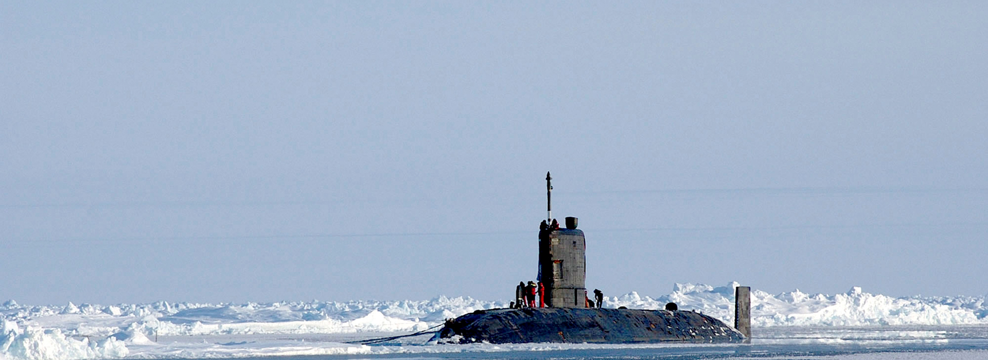 A submarine surfacing through Arctic sea ice, with crew members visible on the conning tower. The vessel is surrounded by broken ice floes against a pale grey winter sky.